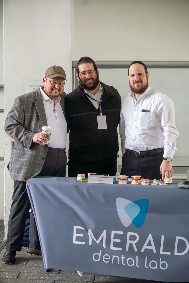 Three attendees posing in front of the Emerald Dental Lab sponsor booth