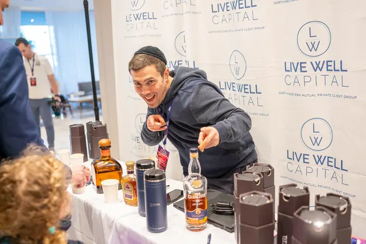 Attendee reaching for items at the LiveWell Capital sponsor table with scotch bottles and promotional items